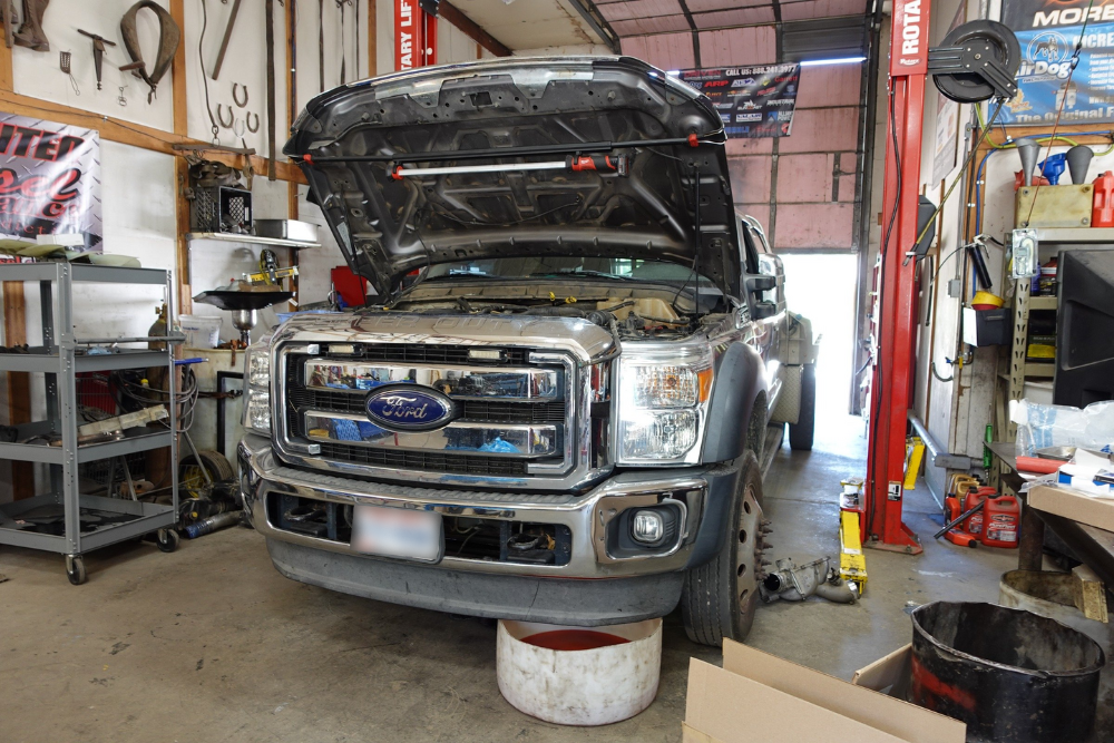 Truck Maintenance Checklist, Diesel Repair Shop in Bremen,OH. Unlimited Diesel Performance. A silver Ford pickup truck sits inside an auto repair shop with its hood open. Tools, shelves, and equipment surround the vehicle, and a drain pan is positioned under the front bumper to catch fluids. The truck is parked between two red vehicle lifts, and the workshop appears busy and well-used.